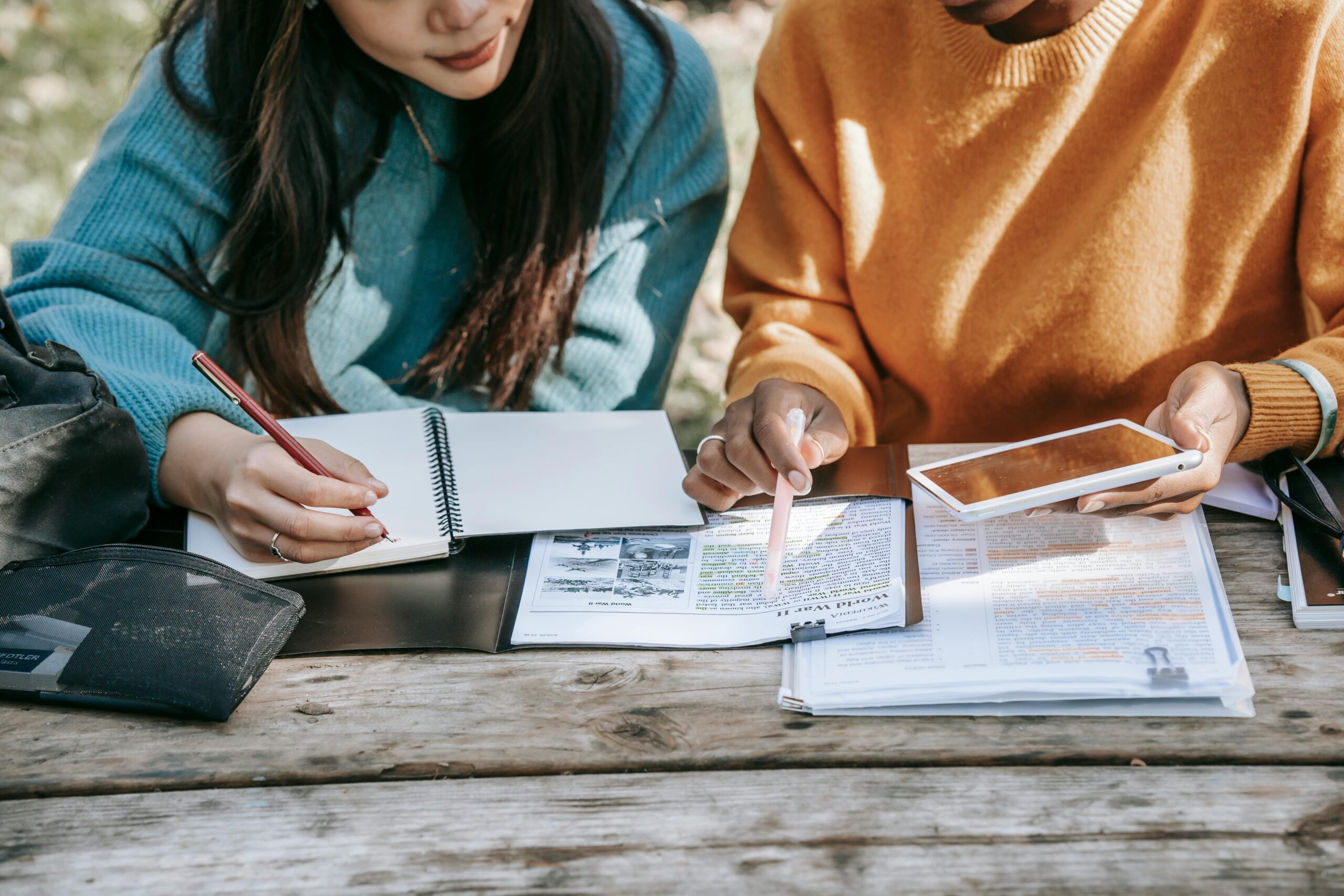 Two students studying together with textbooks and tablets in a park setting.